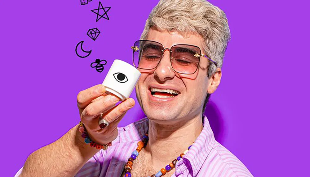 A cheerful person wearing glasses and colorful jewelry holds up a small device with an eye emblem against a purple background with white illustrations floating above