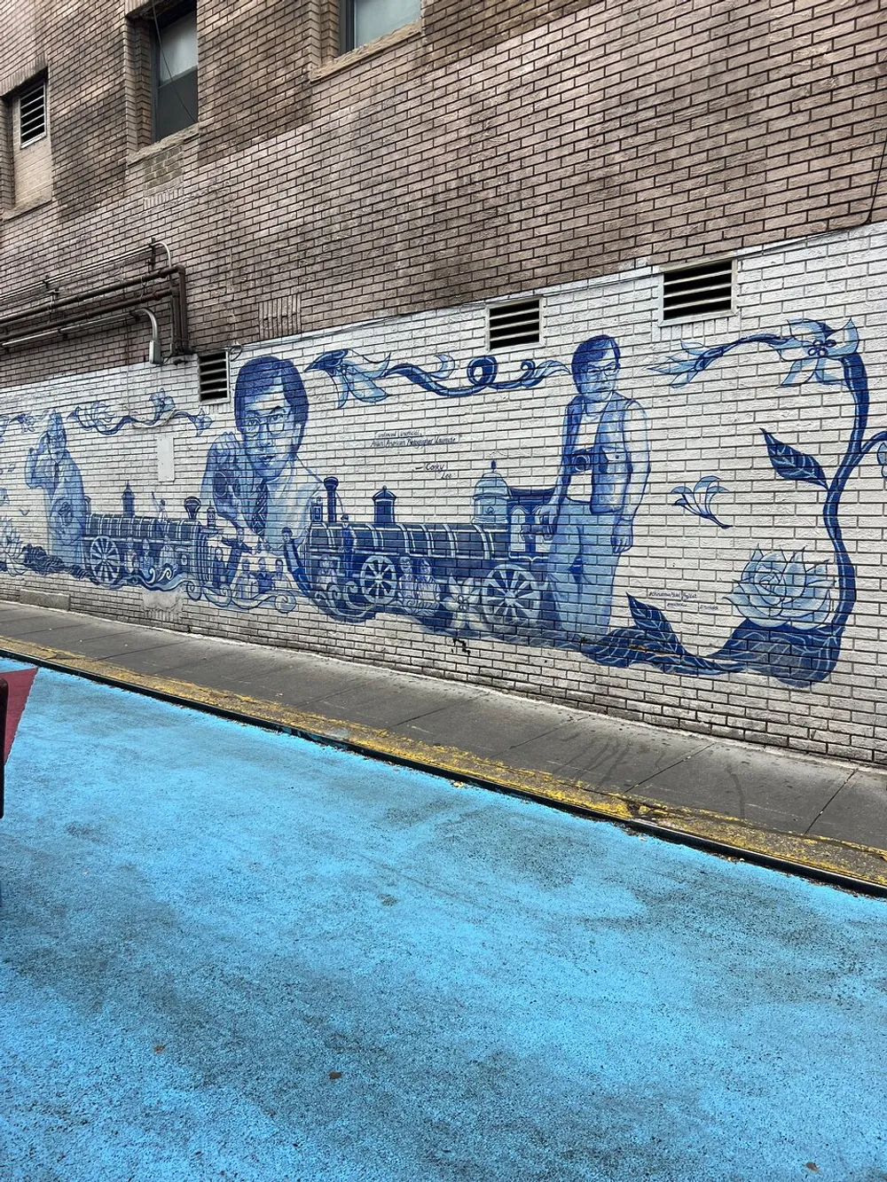 The image shows a detailed blue and white mural on a brick wall depicting individuals and a locomotive complemented by a similarly colored pavement