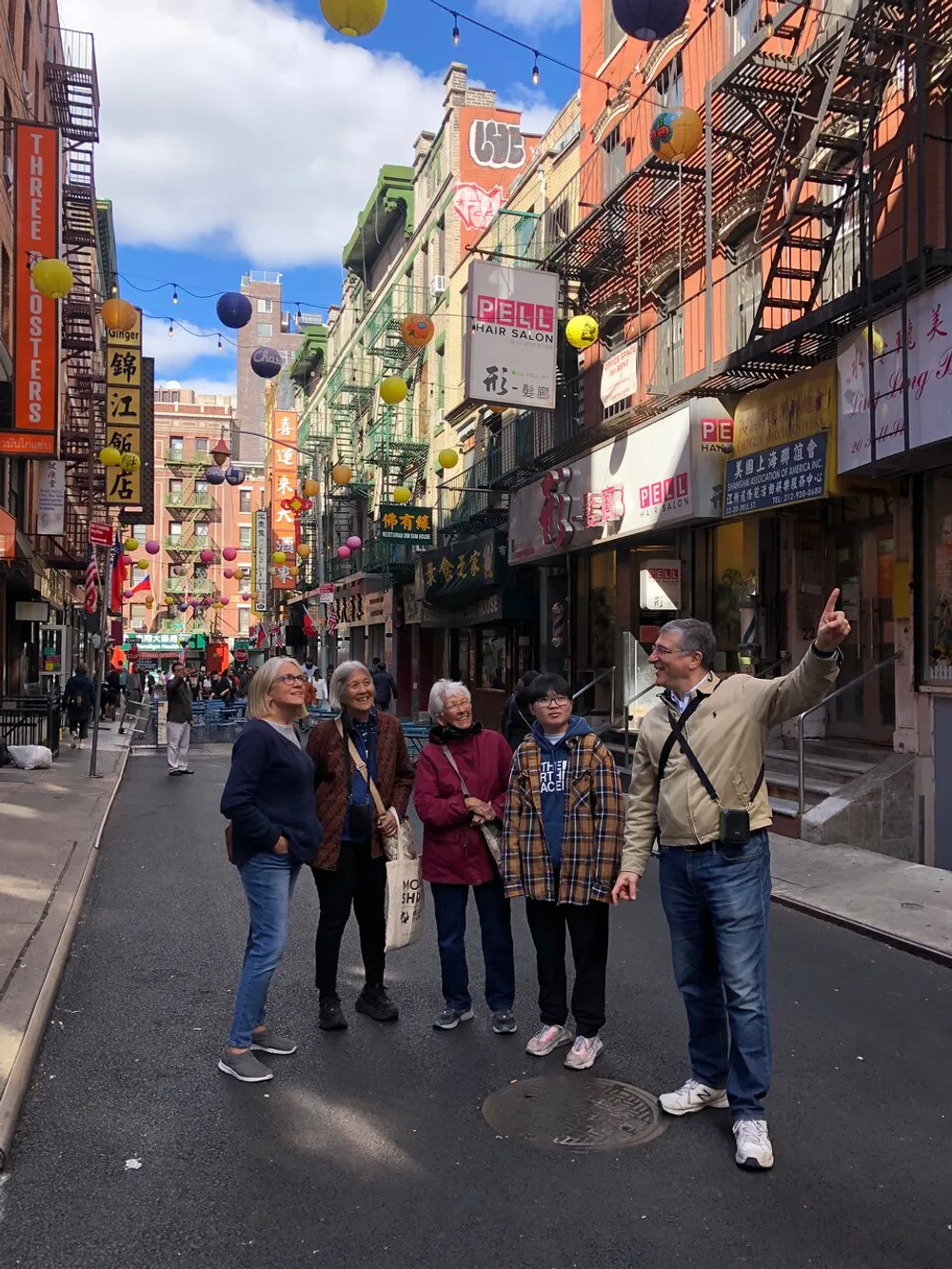A group of five adults appears to be on a tour listening to a man pointing out something of interest in a vibrant street that is likely part of a Chinatown district flanked by colorful signage and traditional lanterns