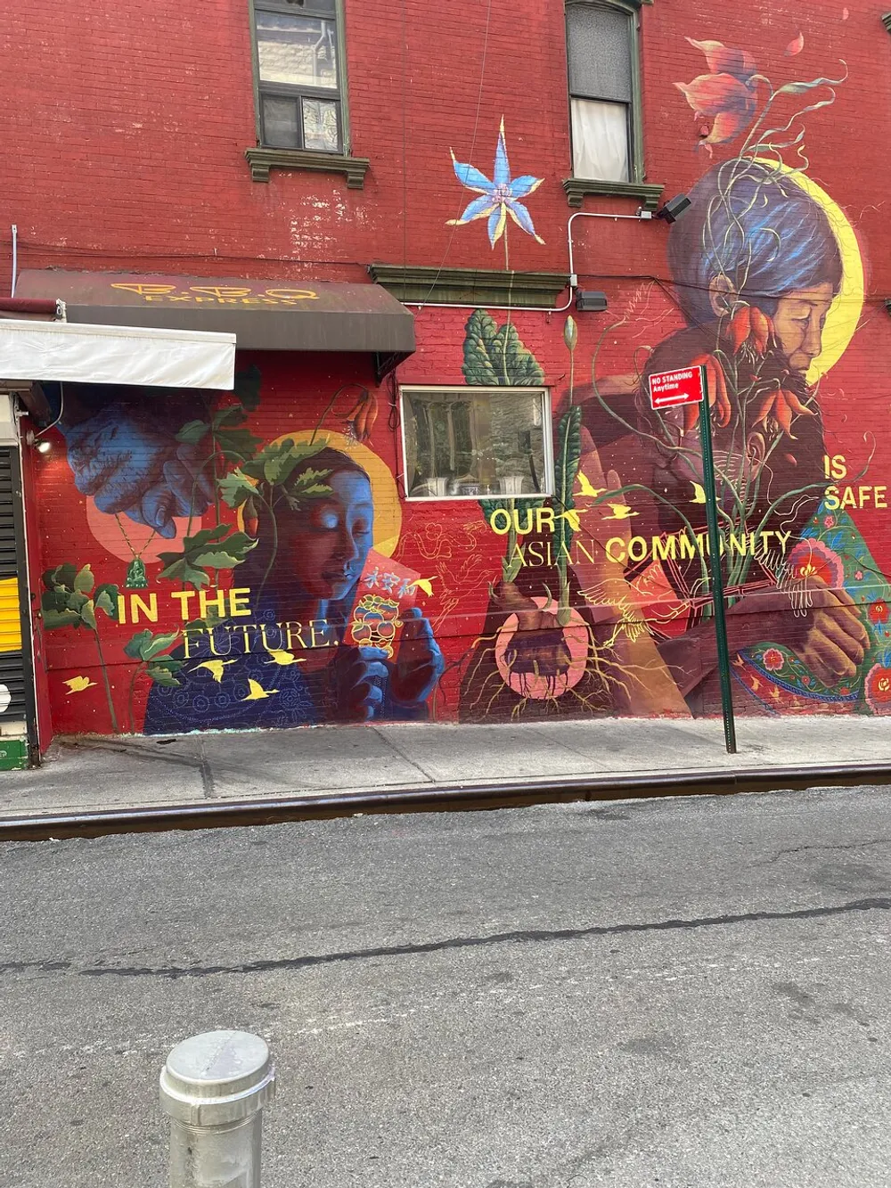 The image shows a vibrant street mural with floral and human imagery and text that reads IN THE FUTURE OUR ASIAN COMMUNITY IS SAFE on the side of a red building