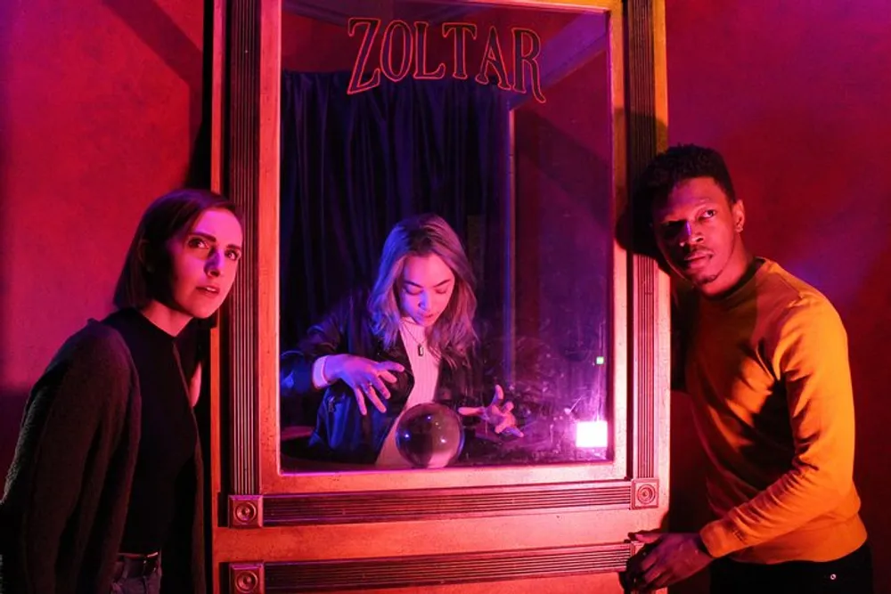 Three individuals are gathered around a Zoltar fortune-telling machine with expressions of intrigue and amusement under ambient red and purple lighting
