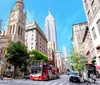 The image showcases a vibrant street view with the iconic Empire State Building towering in the background amidst bright blue skies a red city tour bus in the foreground and bustling urban life
