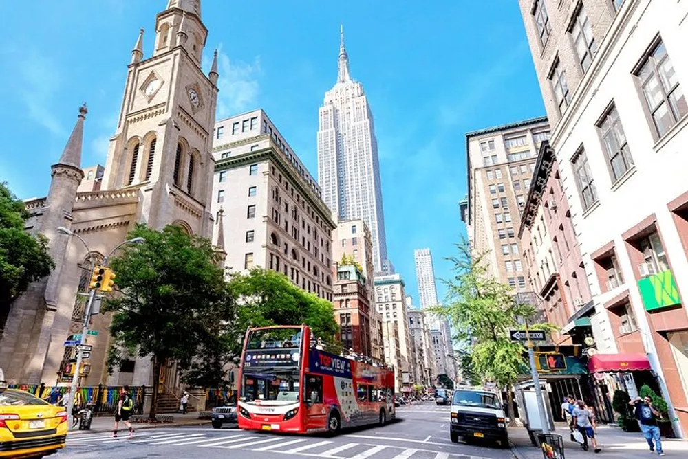 The image showcases a vibrant street view with the iconic Empire State Building towering in the background amidst bright blue skies a red city tour bus in the foreground and bustling urban life