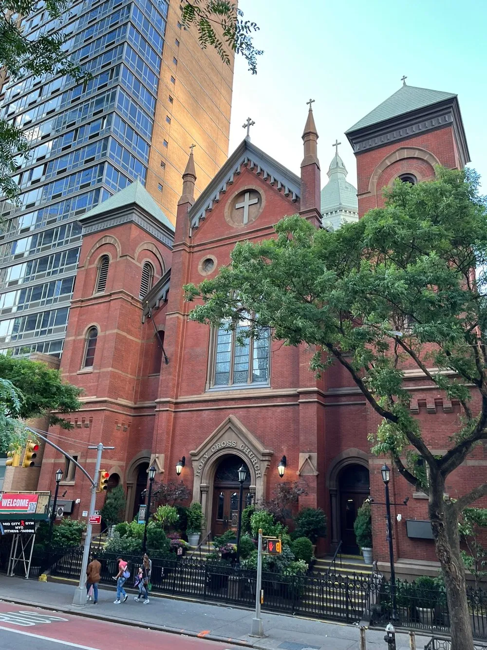 A traditional red brick church with Gothic architectural features nestles among modern buildings and lush green trees under a clear sky