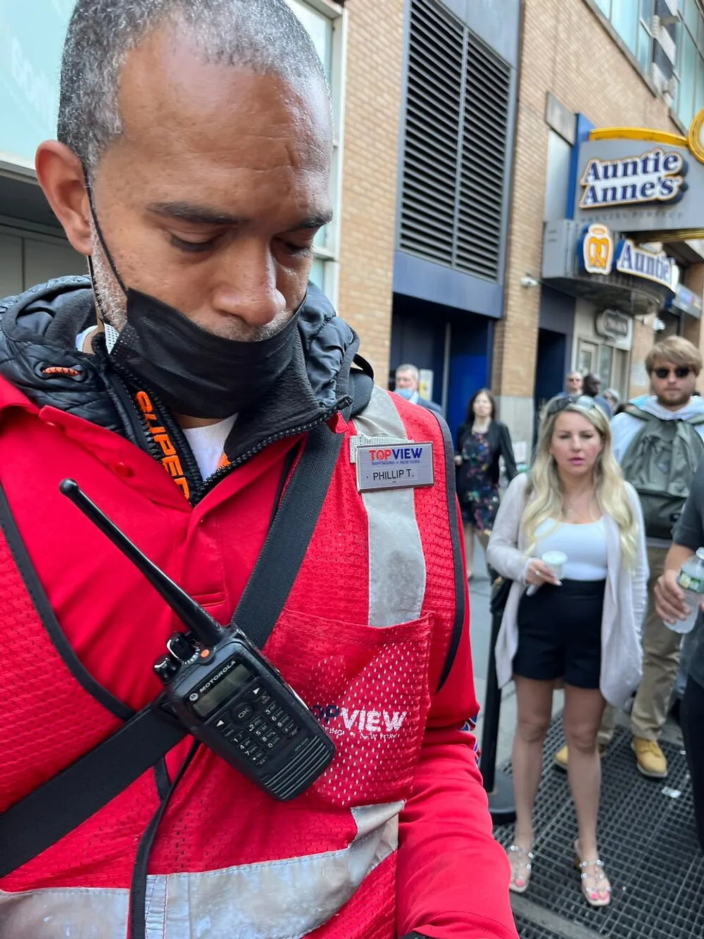 A person wearing a red vest with a TOP VIEW logo looks downwards while a two-way radio hangs on the front and in the background other individuals appear to be walking on a city street