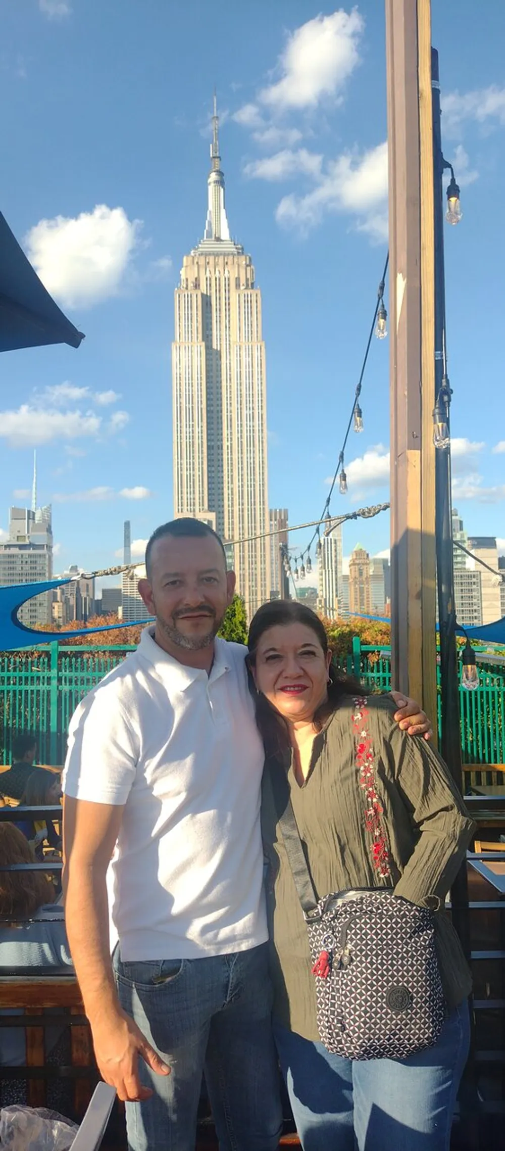 A man and a woman are smiling for a photo with the Empire State Building in the background under a sunny sky