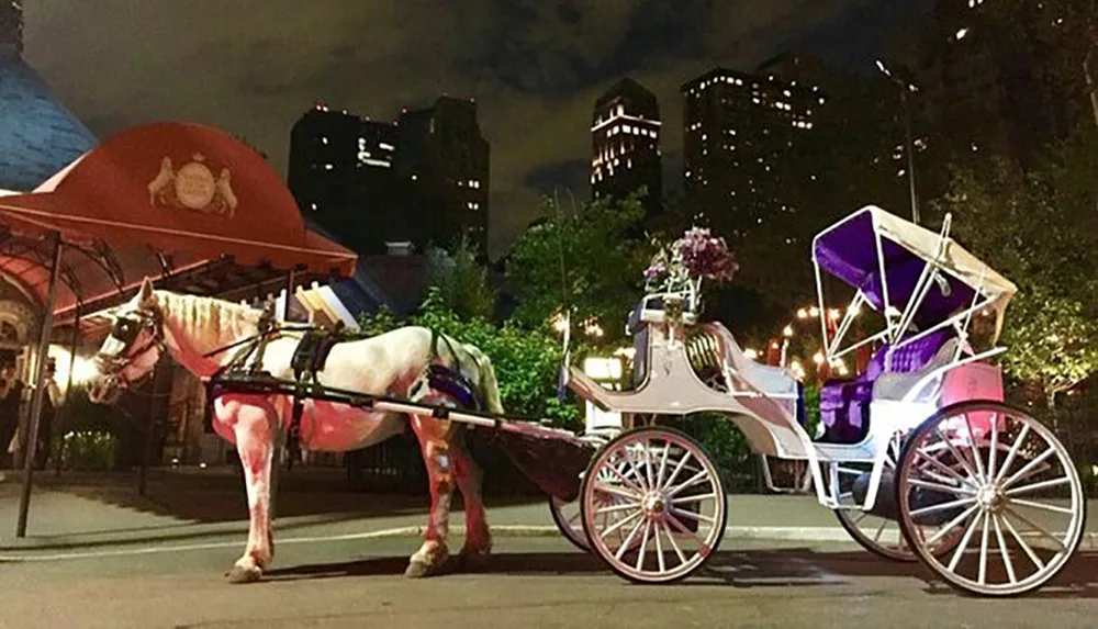 A horse-drawn carriage with pink and white decorations stands at rest against a backdrop of city lights at nighttime