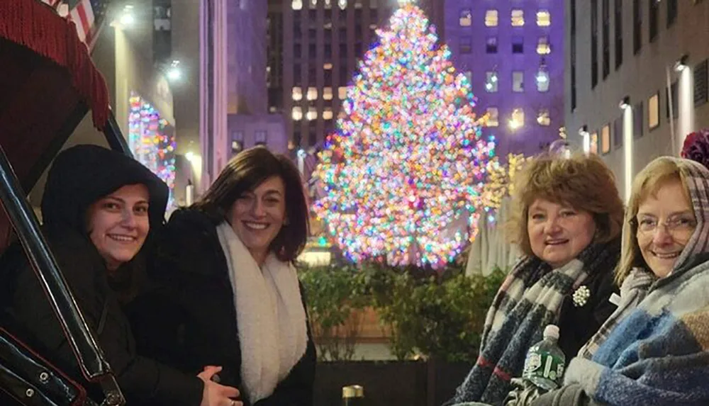 Four people are posing for a photo with a brightly lit Christmas tree in the background likely at a festive outdoor location