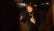 A woman is animatedly speaking outdoors at night, possibly addressing a group, while gesturing with her hands.