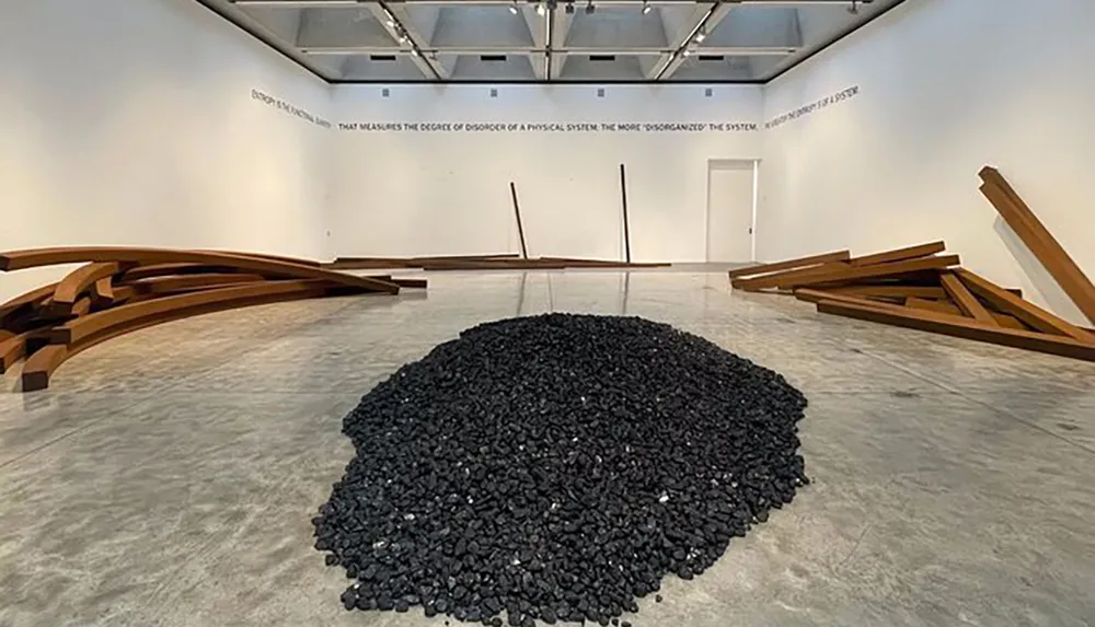 The image shows an art installation with a heap of coal at its center flanked by curved wooden structures within a gallery space accompanied by a text on the wall