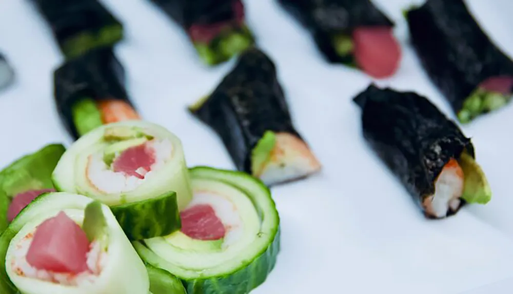 The image showcases a variety of sushi rolls some wrapped in cucumber and others in nori with fillings that include fish and vegetables presented on a white surface
