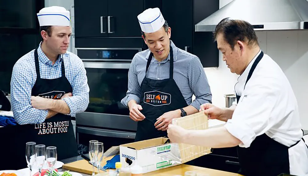 Two men wearing chef hats and aprons watch attentively as a professional chef demonstrates a culinary technique