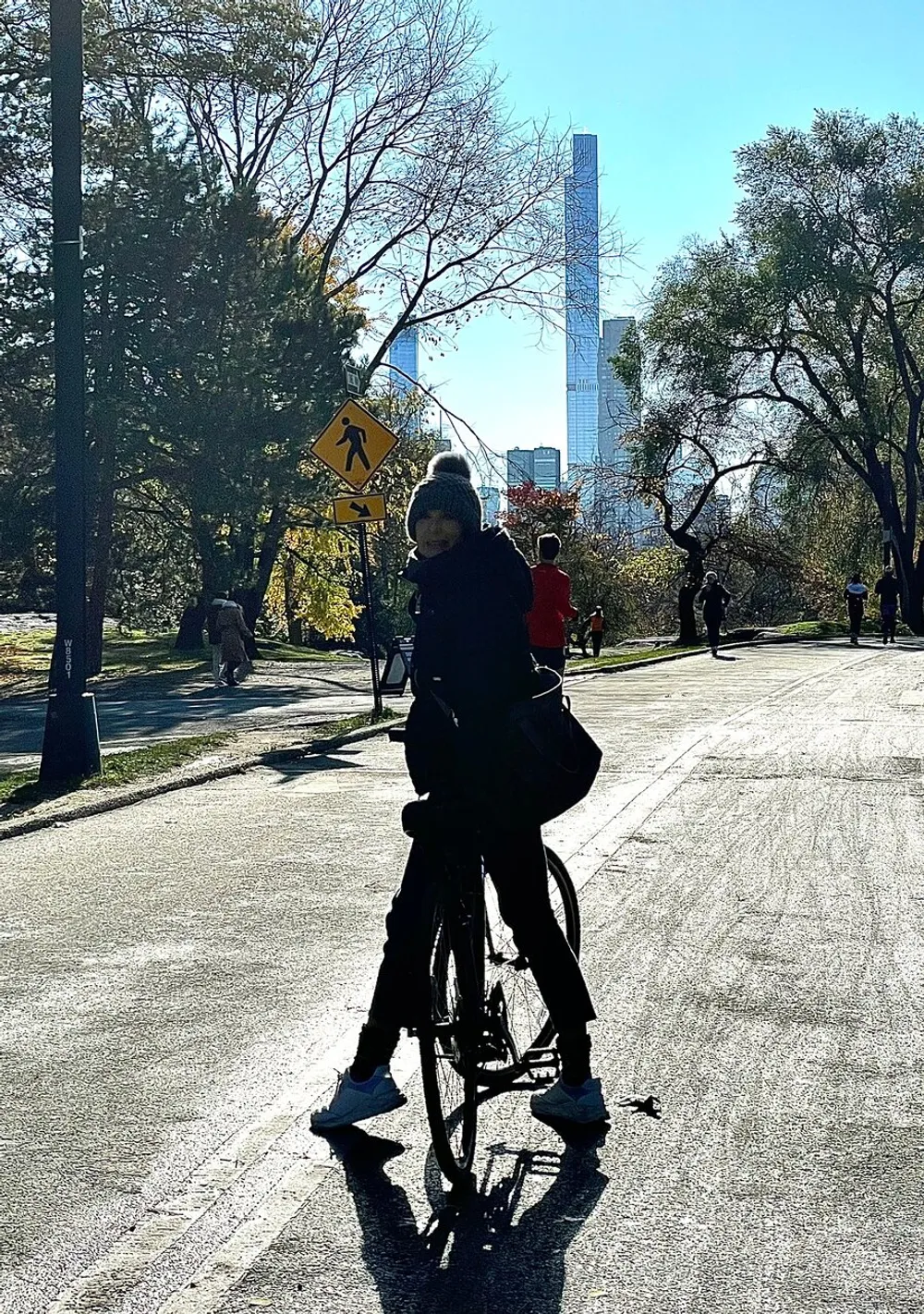 A person on a bicycle is silhouetted against a bright backdrop of trees and skyscrapers in what appears to be an urban park setting