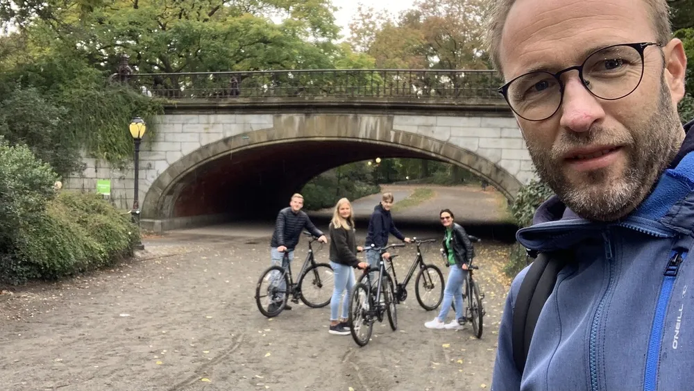 A person is taking a selfie with a group of four people posing with bicycles under a bridge in the background