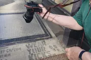 A person is holding a camera next to a dirty doormat with the phrase 