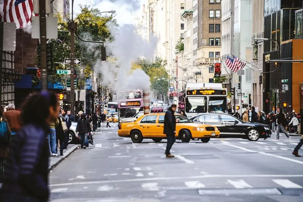A bustling city scene with pedestrians crossing the street a yellow taxi cab in the foreground and steam rising from a street vent