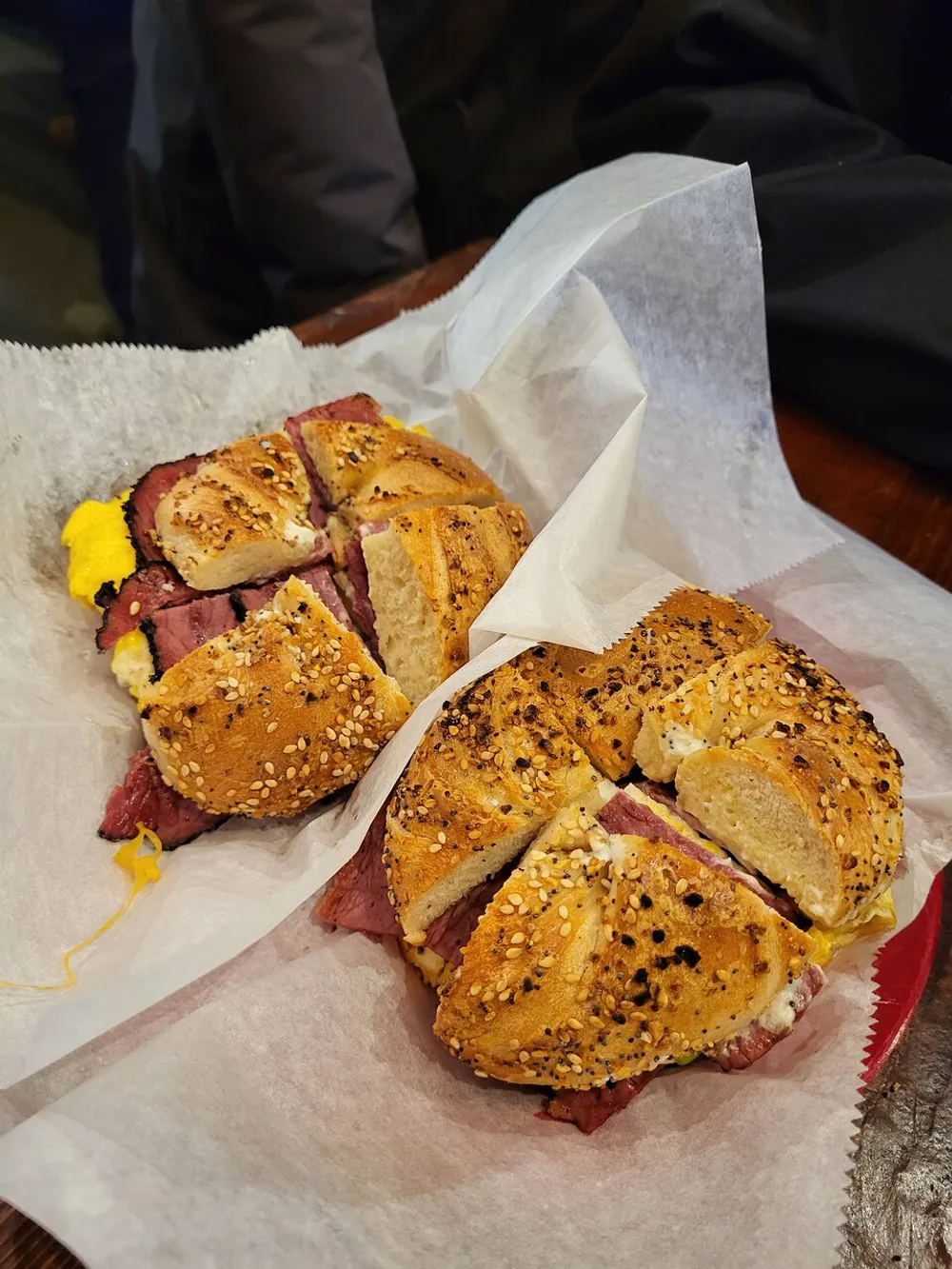 The image showcases a sliced bagel sandwich with beef and mustard on a piece of wax paper with a person partly visible in the background