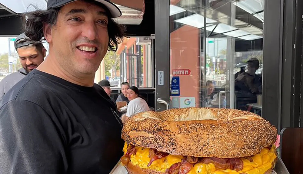 A man is smiling next to an enormous breakfast bagel sandwich that is almost as tall as his torso