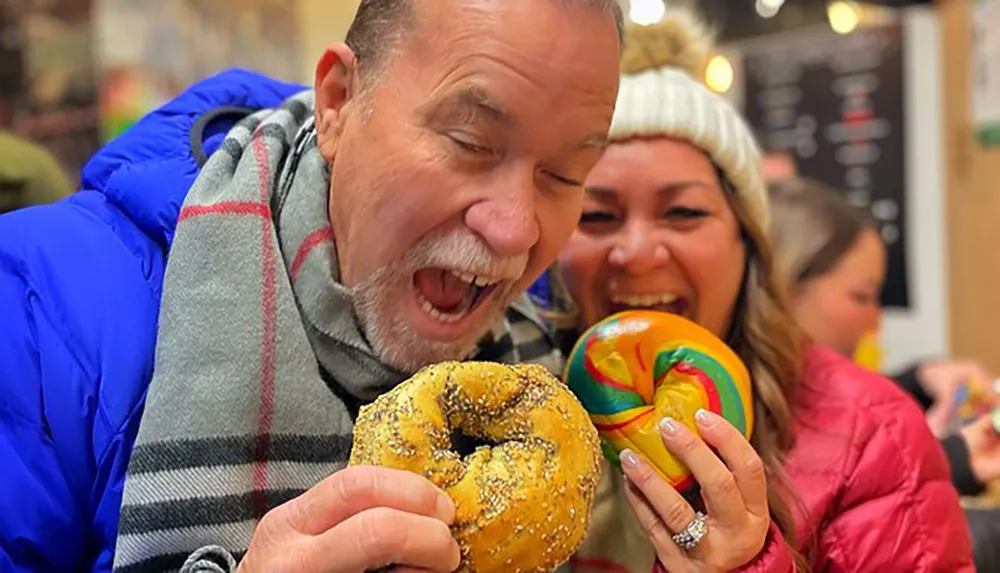 A man and a woman are joyfully pretending to bite into large colorful bagels