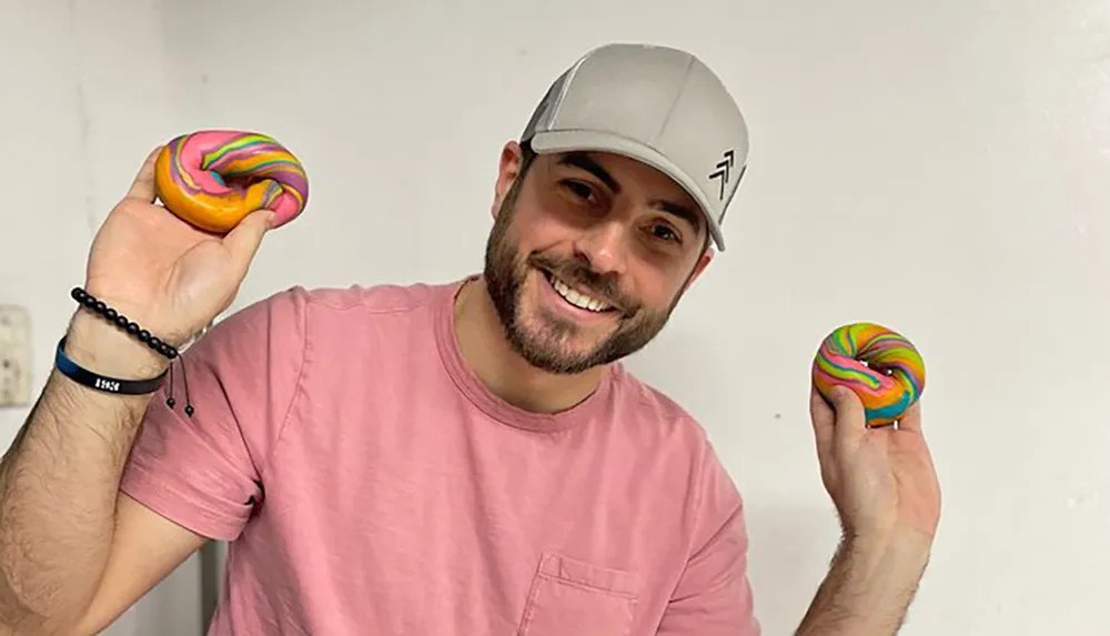 A smiling person in a pink shirt and gray cap holds up two colorfully swirled donuts one in each hand