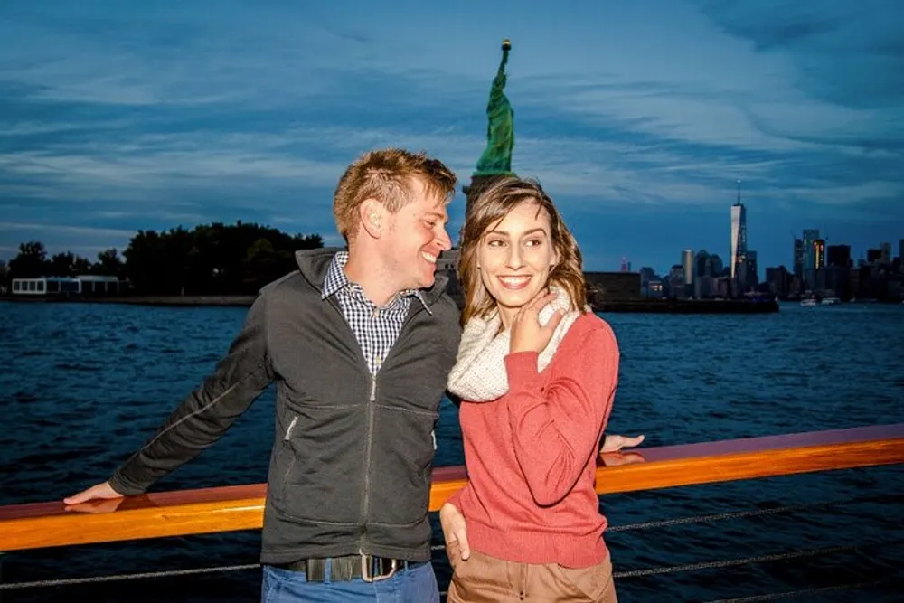 A couple is posing for a photo with the Statue of Liberty and New York City skyline in the background during twilight