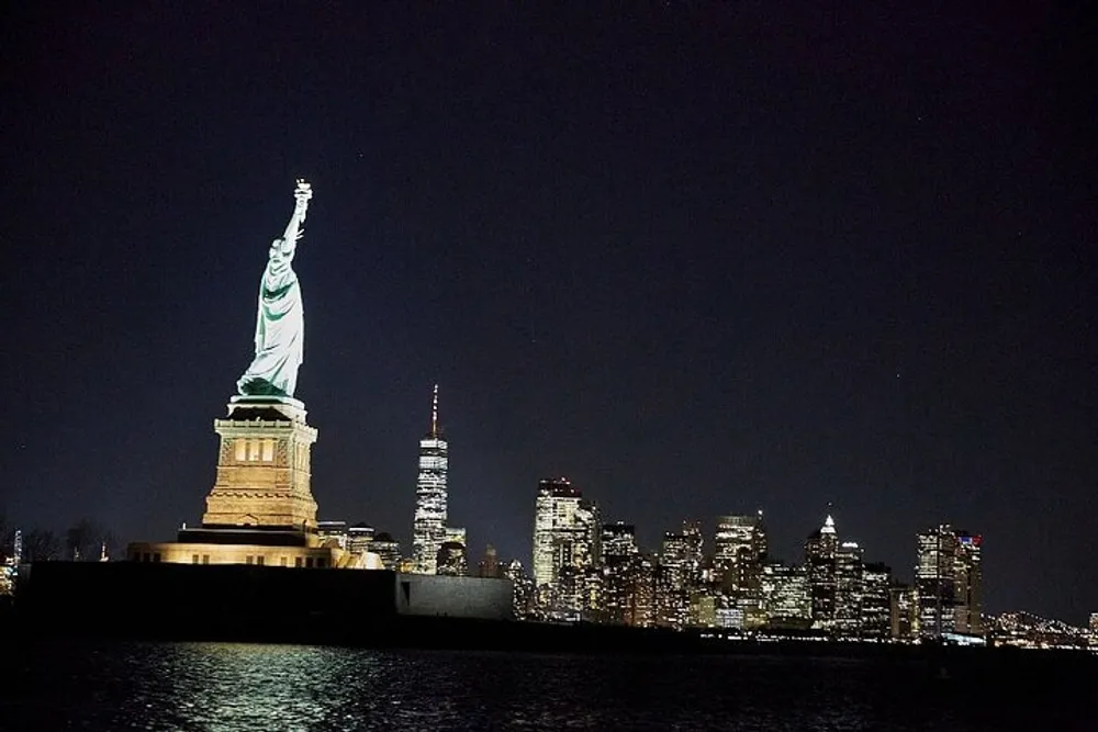 The Statue of Liberty is illuminated at night with the skyline of Lower Manhattan in the background