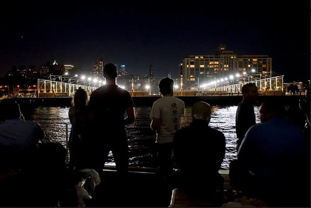 A group of people stands by the water at night looking towards a brightly-lit pier and the urban skyline beyond
