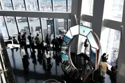 Visitors enjoy a panoramic city view from a high vantage point with large windows, while a person interacts with an installation featuring reflective surfaces.