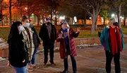A group of people, some wearing masks, are engaged in a conversation or tour outdoors in an urban park at night.