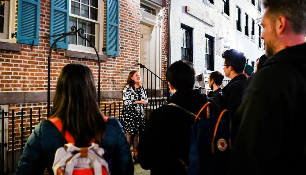 A group of people stand listening to a smiling woman speaking outside a building at night suggesting a guided tour or an outdoor event