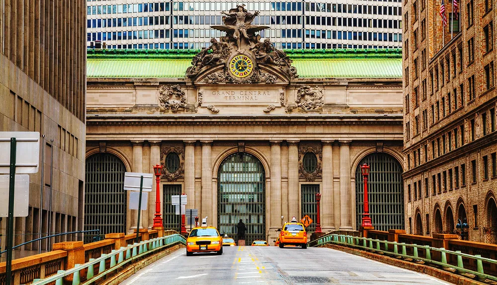 The image shows a street view of the iconic Grand Central Terminal in New York City with yellow taxis in the foreground and the terminals grand facade and ornate statuary clearly visible