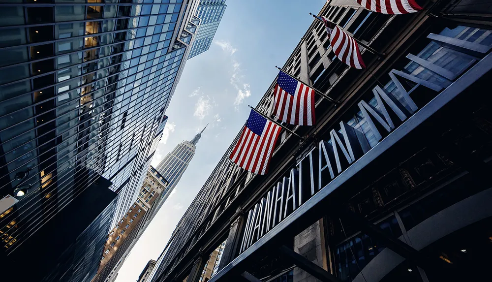 This image captures a view of urban architecture and the Empire State Building framed by American flags flying outside a building with MANHATTAN signage
