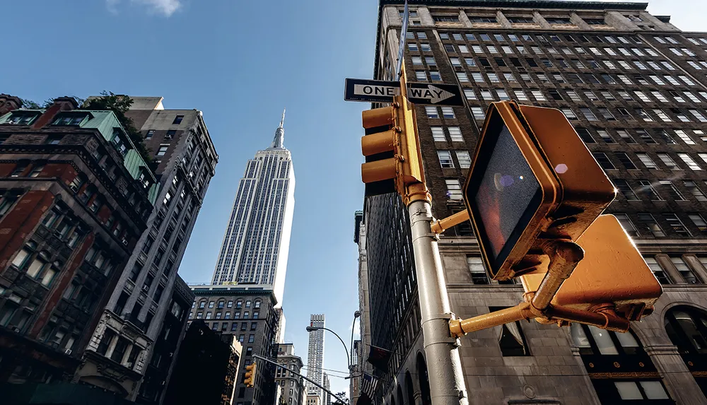 A street view featuring a traffic light and one-way sign with the Empire State Building towering in the background against a clear blue sky