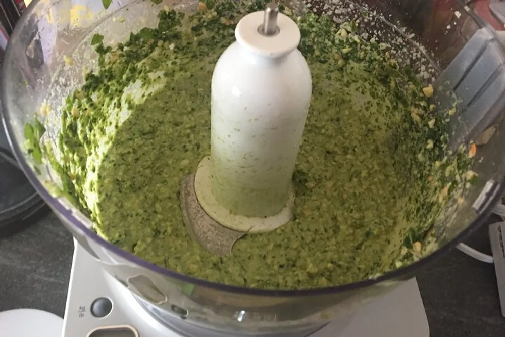 A food processor contains a freshly blended green sauce or mixture likely some kind of pesto or herb-based paste
