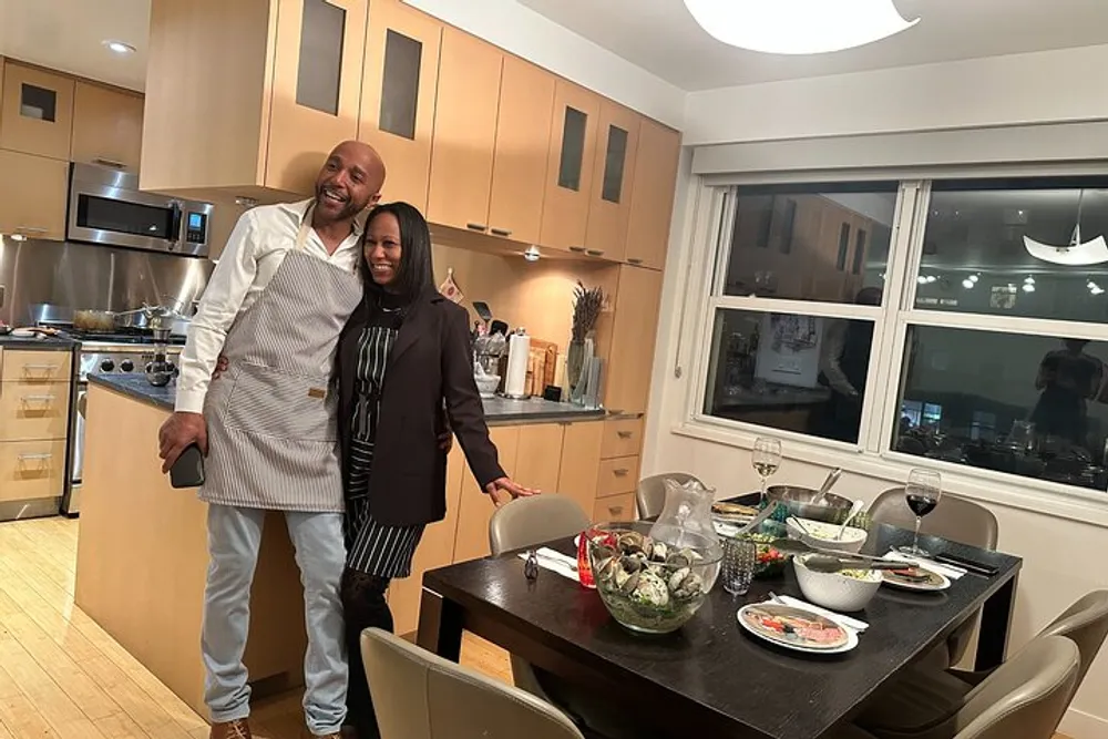 A smiling couple stands in a well-lit modern kitchen with a table bearing dishes of food suggesting a home-cooked meal