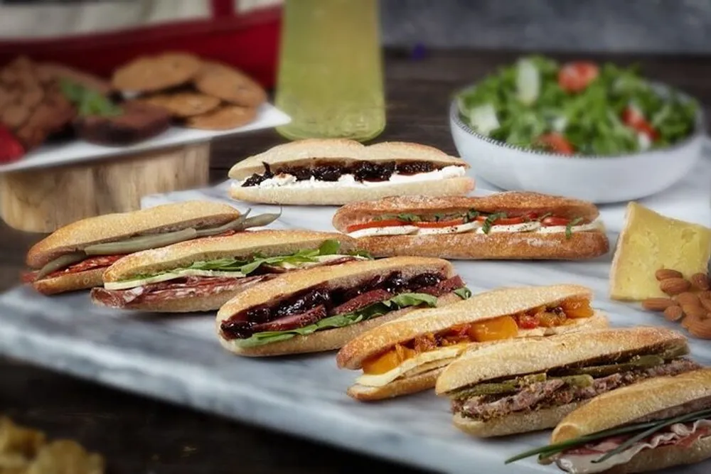 The image features various types of sandwiches possibly with different fillings like meats cheeses and vegetables presented on a striped cutting board with additional food items like salad cookies and a beverage in the background