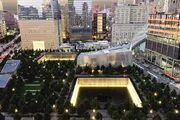 The image shows a high-angle view of the 9/11 Memorial in New York City at dusk, with the illuminated twin reflecting pools surrounded by trees and bordered by modern buildings.