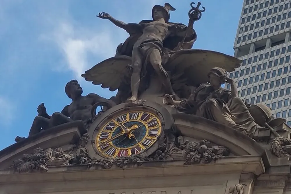 The image shows an ornate clock and sculptures atop the entrance of Grand Central Terminal in New York City against a backdrop of a clear sky and a modern building