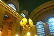 The image features the iconic golden clock in the center of the main concourse of Grand Central Terminal, with its opulent architectural details and soft ambient lighting.