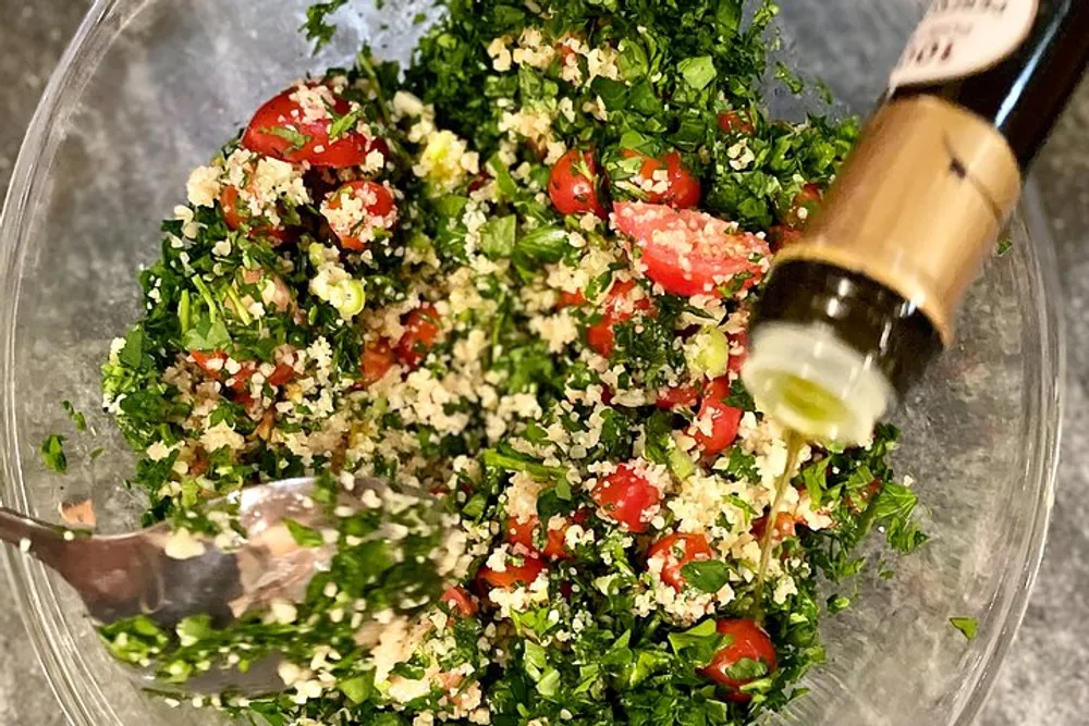The image depicts a glass bowl containing a freshly prepared tabbouleh salad with chopped parsley tomatoes and bulgur and someone is in the process of pouring olive oil into it