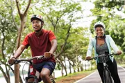 Two people wearing helmets are riding bicycles along a tree-lined path.