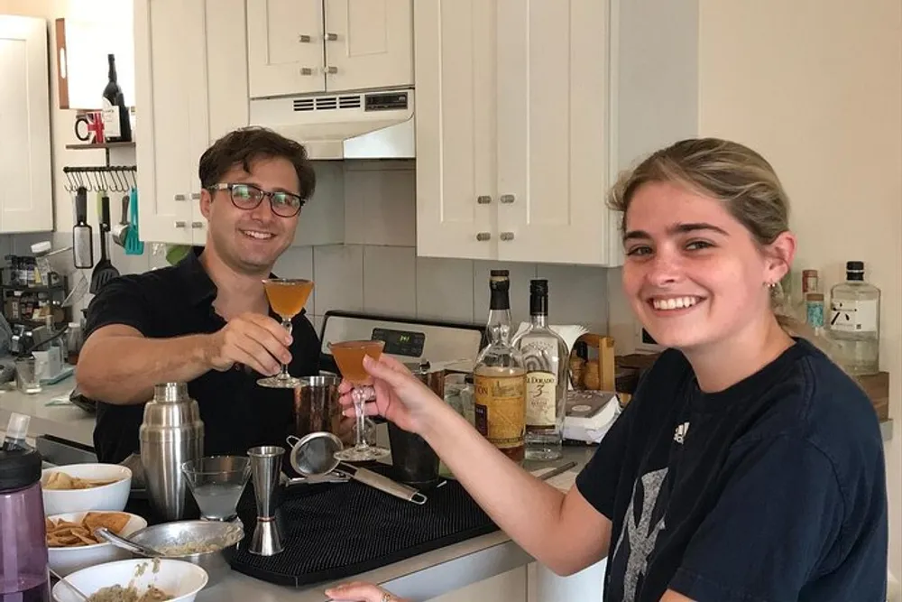 Two people are cheerfully toasting with cocktails in a home kitchen setting
