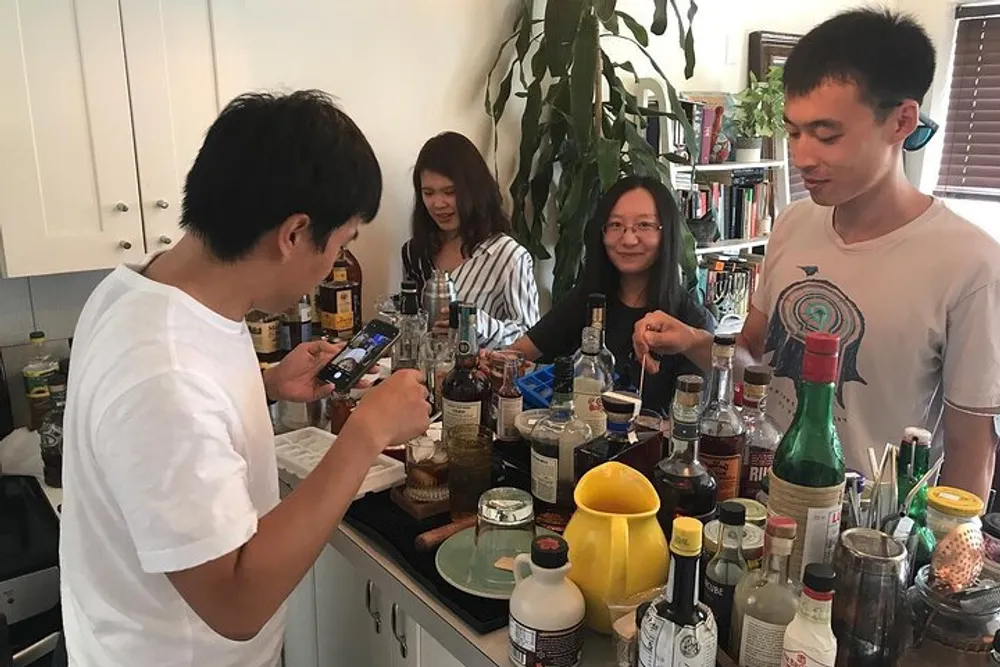 Three individuals are socializing in a kitchen with one person taking a photo while surrounded by a variety of bottles likely containing alcoholic beverages