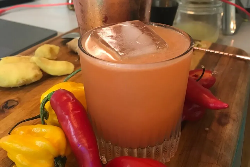 A glass of a pinkish-orange drink with ice is placed on a wooden board surrounded by colorful chili peppers and ginger slices