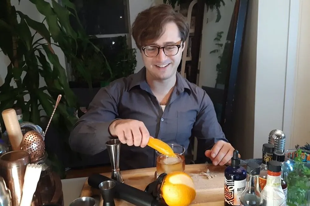 A smiling person is garnishing a drink with an orange peel at a home bar setup surrounded by various cocktail-making tools and ingredients