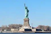 The image shows the Statue of Liberty under a clear blue sky, seen from a distance with water in the foreground.