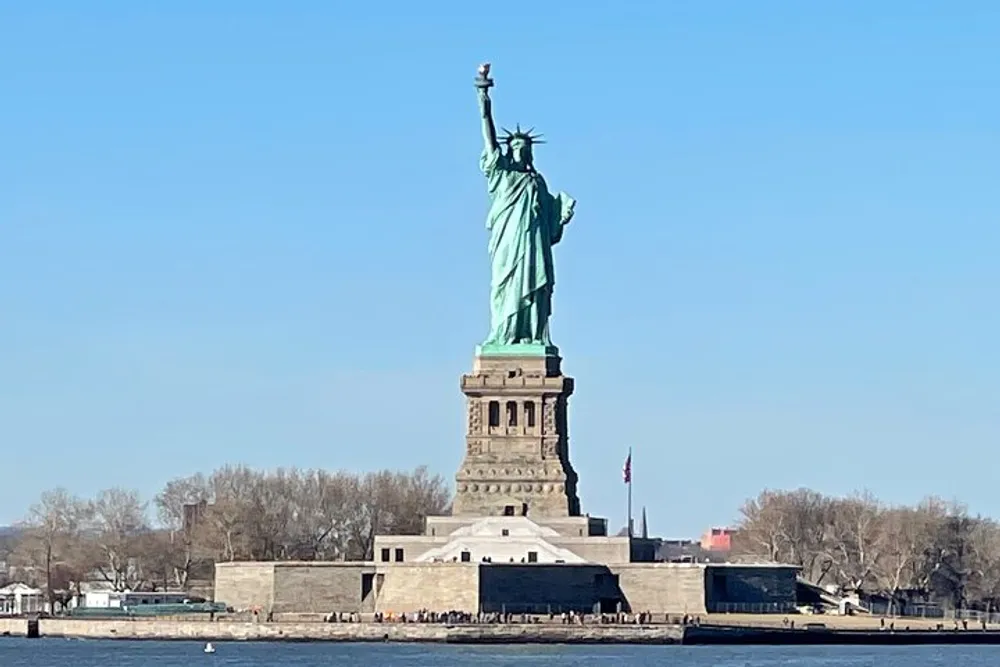 The image shows the Statue of Liberty under a clear blue sky seen from a distance with water in the foreground