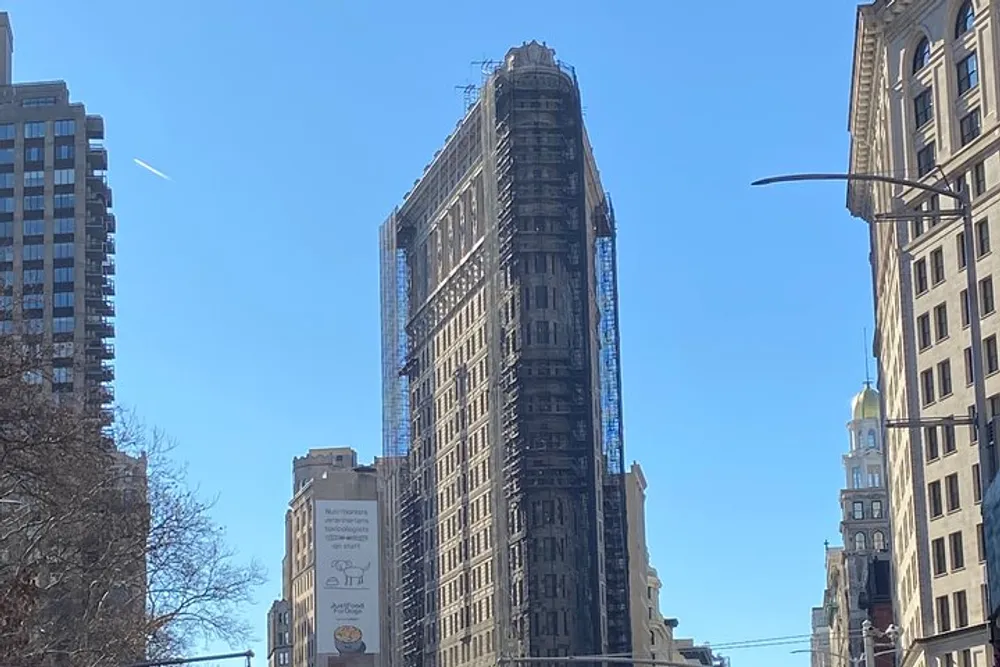 The image shows a flatiron-shaped skyscraper covered in scaffolding under a clear blue sky in an urban setting