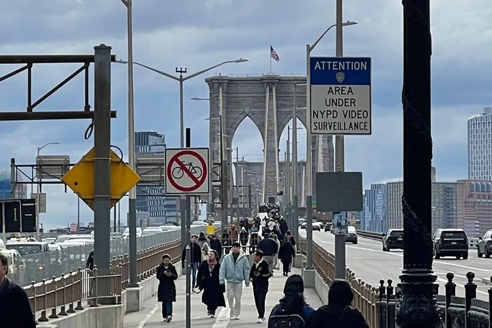 Pedestrians walk along the footpath of a bridge under NYPD video surveillance on a cloudy day with an iconic bridge in the background