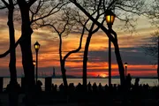 Silhouetted figures and bare trees are set against a vivid sunset sky, with the glow of street lamps adding to the tranquil evening ambiance by the water.